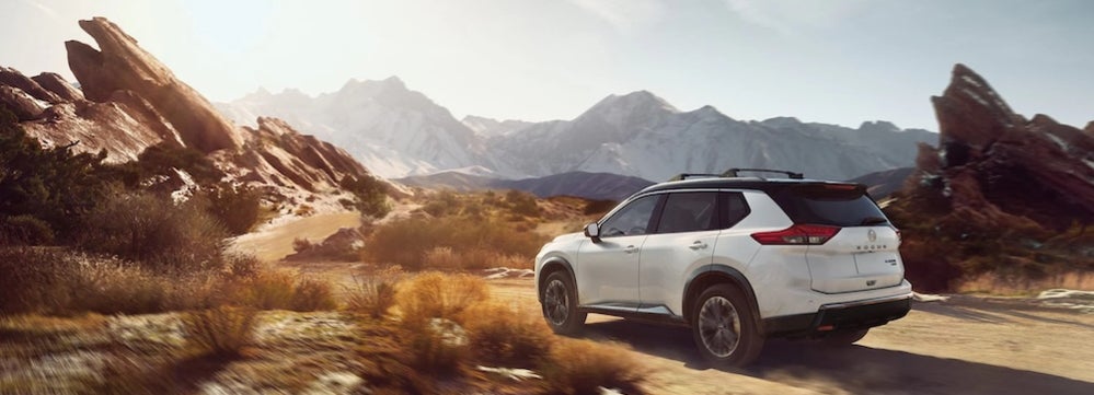 A white 2026 Nissan Rogue SUV driving through desert scrubland toward a sharp outcropping of red rocksALT