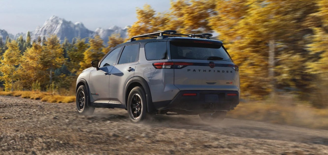 A gray 2026 Nissan Pathfinder SUV driving down a dirt road with yellow trees and mountains in the background