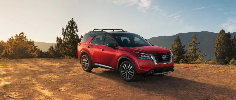 A three-quarters view of a red 2025 Nissan Pathfinder SUV parked on a bare dirt patch, with mountains in the background